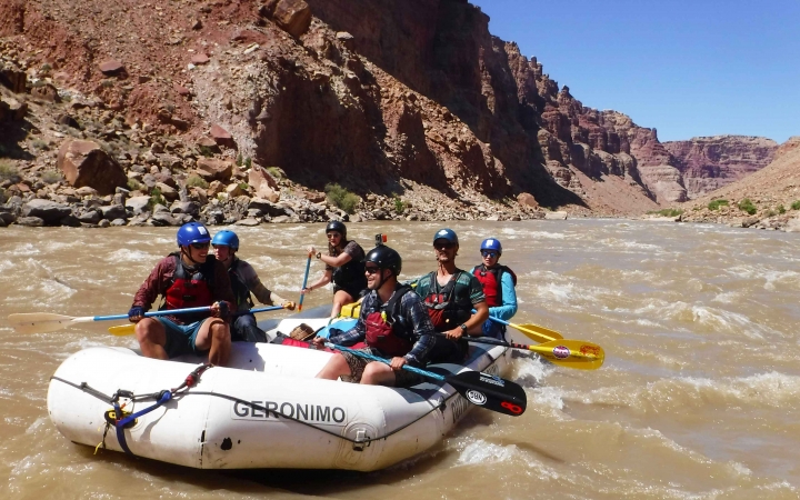 A group of students wearing safety gear paddle a raft on a choppy river. The river is framed by high canyon walls.